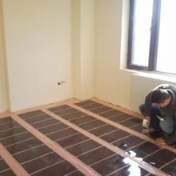Instalacja mat grzewczych na podczerwień na podłodze w jasnym pomieszczeniu; monter przykleja maty taśmą klejącą wzdłuż krawędzi.