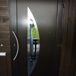 Nowoczesne, brązowe drzwi wejściowe z unikalnym, łezkowatym przeszkleniem, zamontowane w drewnianej elewacji domu. Widoczny srebrny uchwyt i wycieraczka.