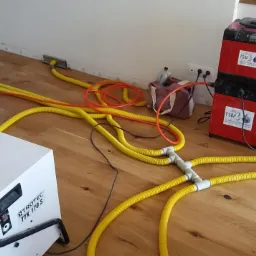 Dwa urządzenia do osuszania, czerwone i białe, połączone żółtymi wężami na drewnianej podłodze, z widocznymi gniazdkami elektrycznymi w ścianie.