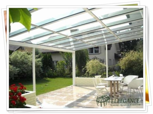 Biała pergola z przezroczystym dachem na tarasie z marmurową posadzką, widok na zadbany ogród i dom. Stolik z krzesłami pod pergolą.