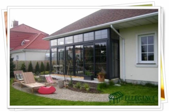 Szklana zabudowa tarasu w ciemnej ramie, z widokiem na ogród z leżakami i ścieżką z kamienia. Dom z czerwoną dachówką w tle.