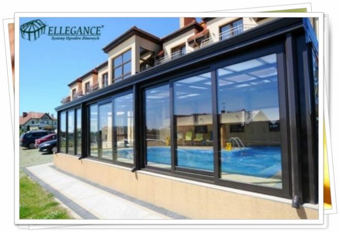 Przeszklona zabudowa tarasu z widokiem na basen. Czarna konstrukcja, duże okna przesuwne. W tle budynek z balkonami i fragment parkingu. Błękitne niebo, słoneczny dzień.