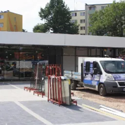 Szklane panele w metalowych stojakach przed budynkiem z przeszkloną fasadą, obok zaparkowany samochód dostawczy z logo firmy, w tle budynki mieszkalne.