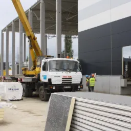 Budowa hali z użyciem dźwigu Tatra i płyt warstwowych Kingspan, widoczni pracownicy w odblaskowych kamizelkach i kaskach.