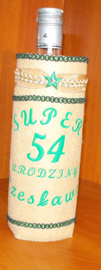 Ozdobna butelka na alkohol z haftowanym napisem 'Super 54 Urodziny Czesława' i dekoracjami z zielonych koralików oraz wstążki ze złotą gwiazdą, umieszczona na drewnianej półce.