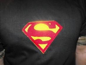 Czarna koszulka z wyhaftowanym, czerwono-żółtym logo Supermana na plecach, widoczny fragment rękawa w kratę. Detal haftu.