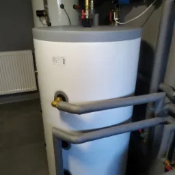 Biały zbiornik akumulacyjny ciepłej wody użytkowej z szarym wykończeniem, widoczne rury z izolacją termiczną oraz zawory, na tle grzejnika panelowego i szarej ściany.