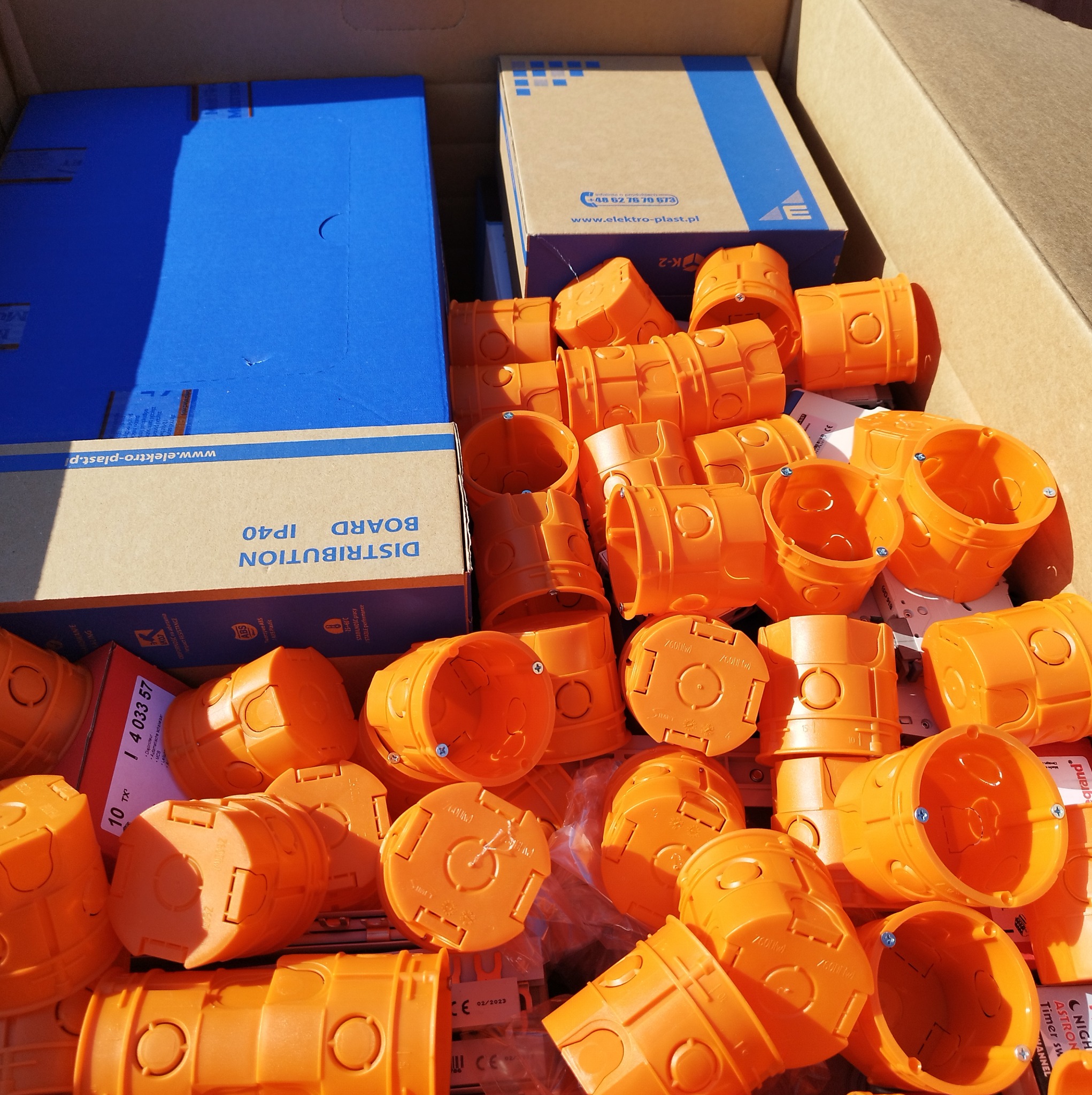 Pomarańczowe puszki instalacyjne elektryczne, wraz z opakowaniami producenta Elektro-Plast, zgromadzone w kartonowym pudle w Mogilnie.