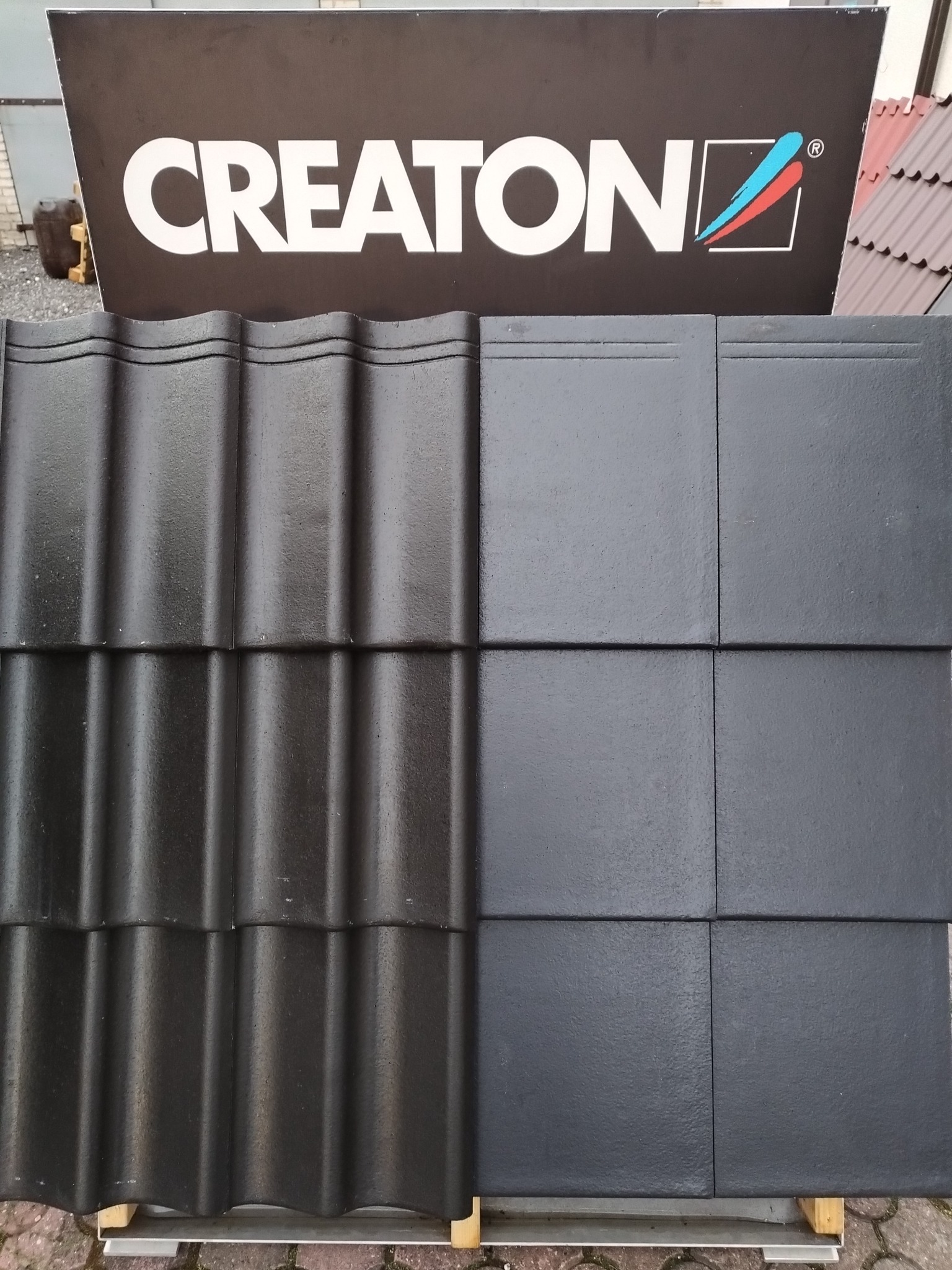 Ekspozycja dwóch rodzajów czarnej dachówki CREATON: falistej i płaskiej, prezentowanych pod tablicą z logo producenta.