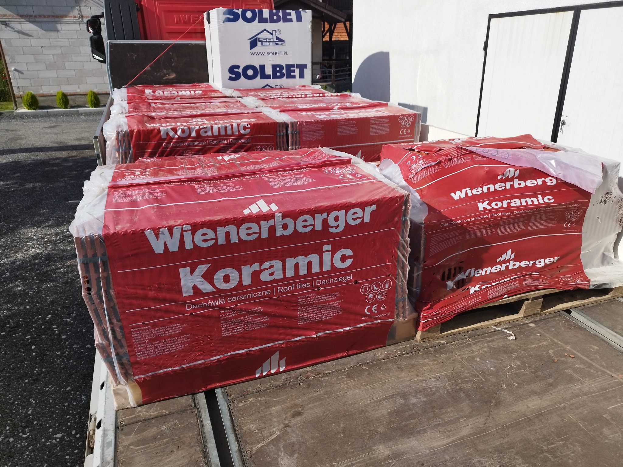 Palety z czerwoną dachówką ceramiczną Wienerberger Koramic, zabezpieczone folią, ułożone na pace samochodu dostawczego. W tle widoczny biały blok Solbet.