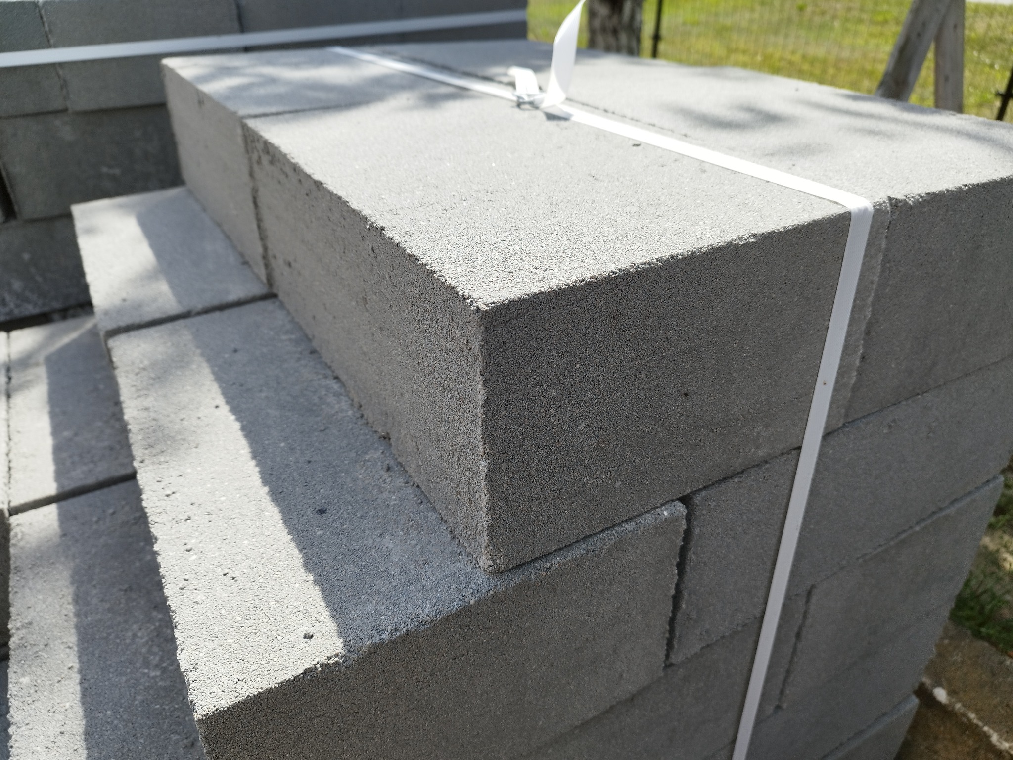 Szare bloczki betonowe ułożone w stos, zabezpieczone białą taśmą, widoczne na zewnątrz w świetle słonecznym.