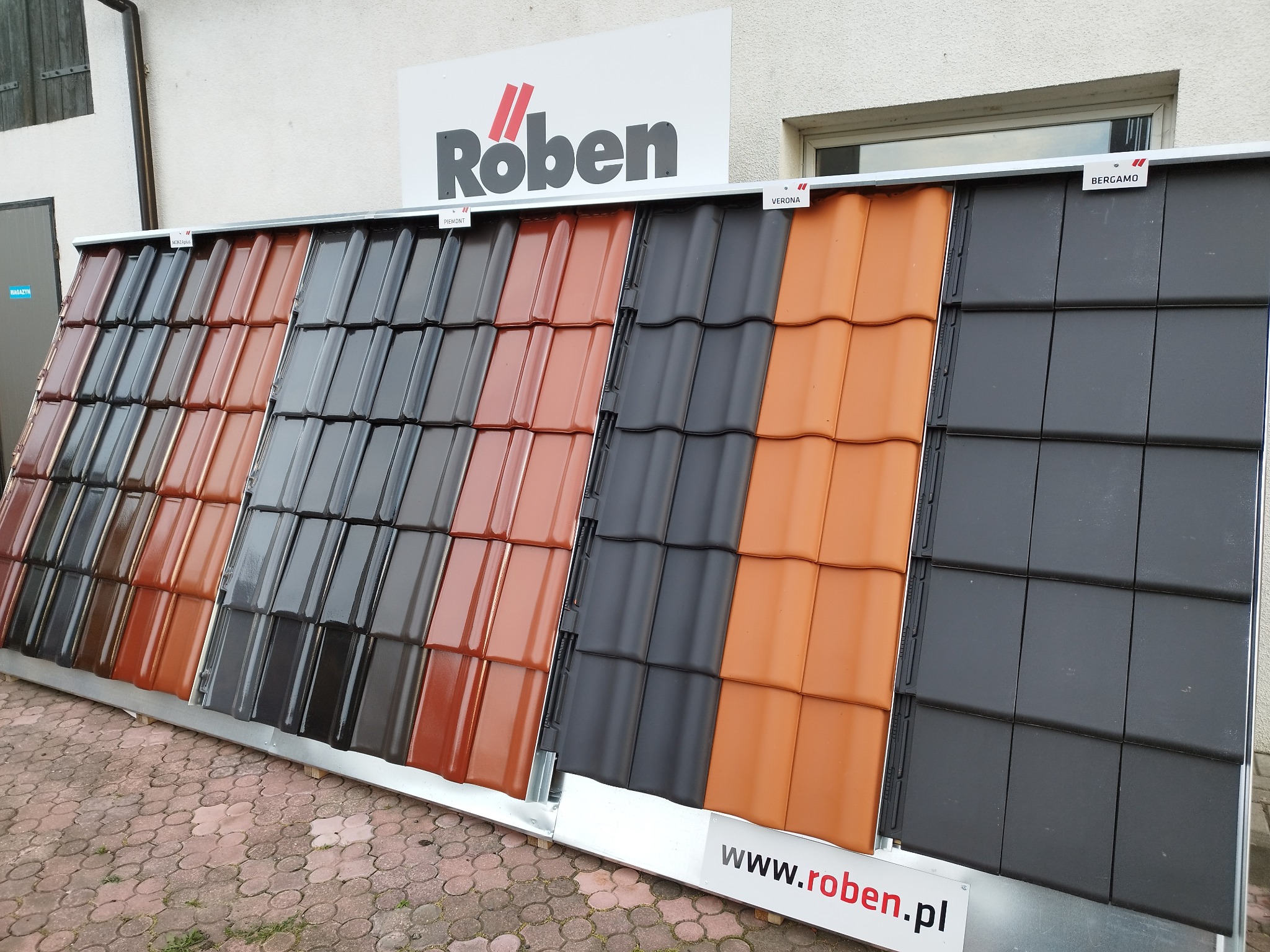 Ekspozycja dachówek Roben w różnych kolorach i modelach (Piemont, Verona, Bergamo) na zewnątrz budynku, z widocznym logo producenta i adresem strony internetowej.