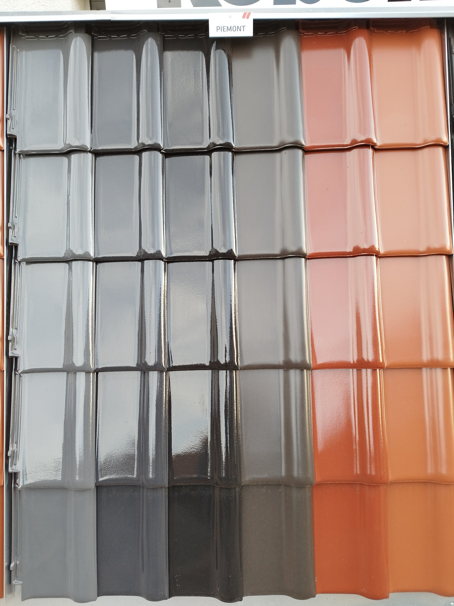 Różnokolorowe dachówki ceramiczne marki Piemont, prezentowane obok siebie w ekspozytorze, odcienie szarości i brązu.