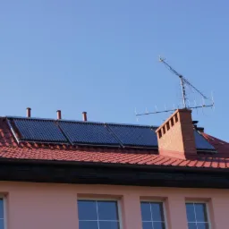 Czerwony dach budynku z ceglanymi kominami i panelami solarnymi pod błękitnym niebem. Widoczne okna budynku.