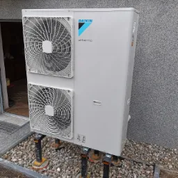 Zewnętrzna jednostka pompy ciepła Daikin Altherma, zamontowana na metalowych wspornikach na żwirowym podłożu przy szarej elewacji budynku.