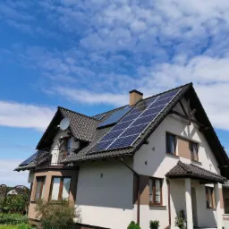 Dom jednorodzinny z panelami fotowoltaicznymi na dachu, widok z dołu na tle błękitnego nieba z chmurami.