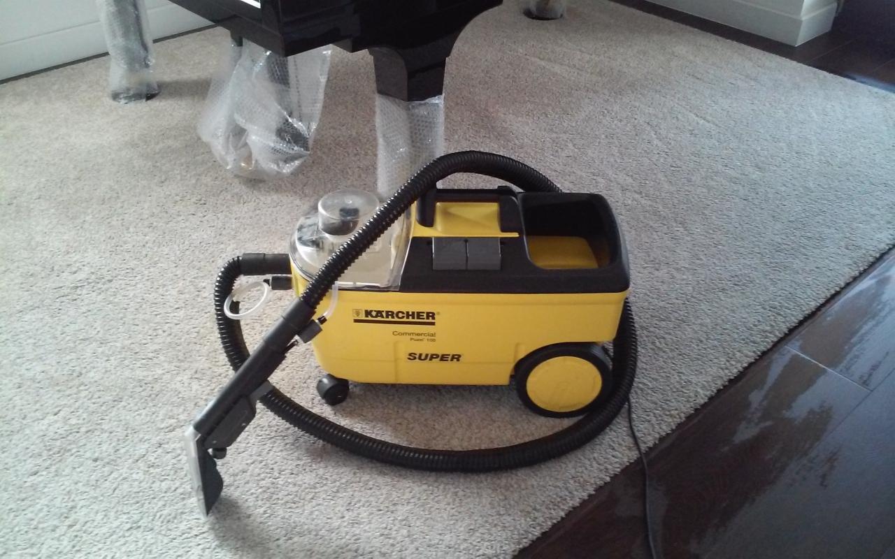 Żółty odkurzacz piorący Karcher na dywanie z długim włosiem, widoczne nogi mebli zabezpieczone folią. Sprzęt do czyszczenia dywanów.