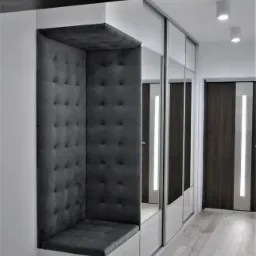 Wnęka z siedziskiem obitym szarą, pikowaną tkaniną, wkomponowana w ciąg szafy przesuwnej z lustrzanymi frontami w jasnym przedpokoju.