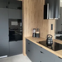 Nowoczesna kuchnia: szare, matowe fronty szafek z drewnianym blatem, czarny czajnik na płycie indukcyjnej, blender i nóż w bloku na blacie, czarny okap cylindryczny.