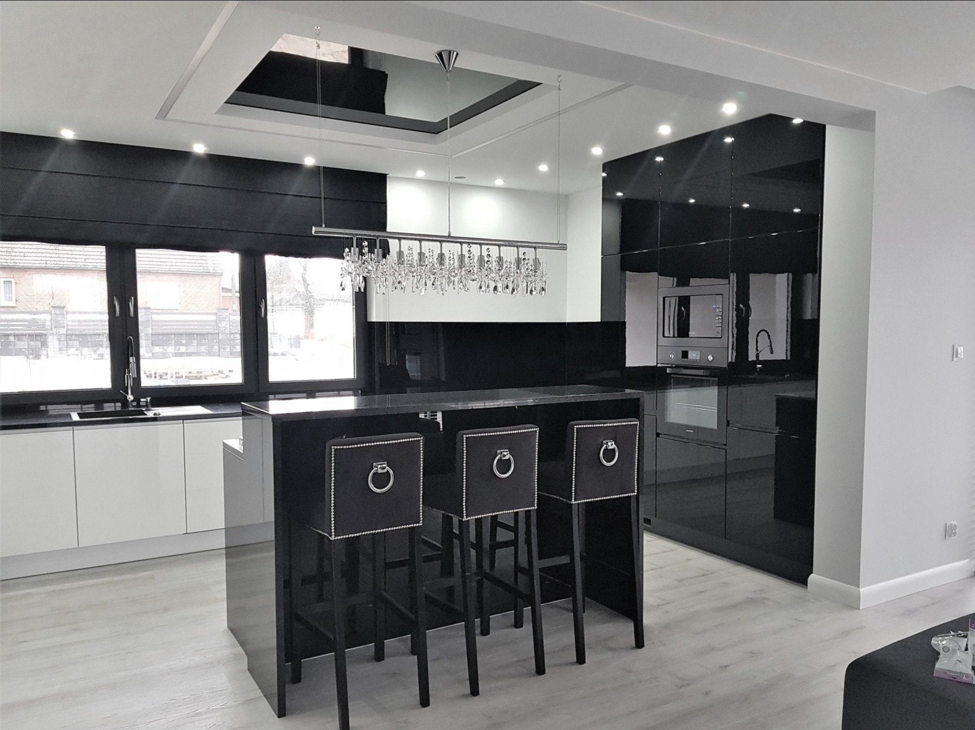 Nowoczesna kuchnia w czerni i bieli z wyspą, trzema hokerami z ozdobnymi uchwytami, kryształowym żyrandolem i czarnymi roletami rzymskimi w oknie.