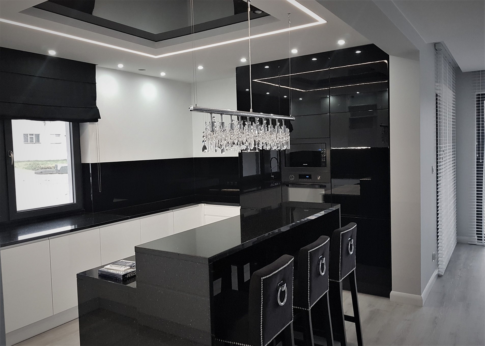 Nowoczesna kuchnia w czerni i bieli z wyspą, oświetleniem LED w suficie i kryształowym żyrandolem nad blatem. Czarne, lakierowane fronty szafek kontrastują z białymi elementami, a wysokie krzesła...
