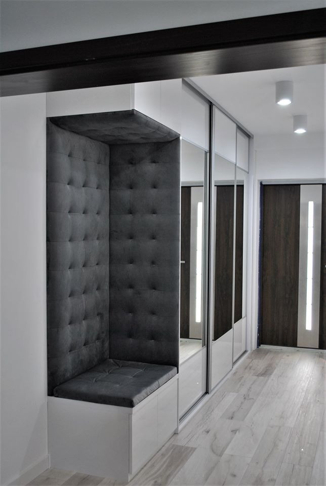 Wnęka z siedziskiem obitym szarą, pikowaną tkaniną, wkomponowana w ciąg szafy przesuwnej z lustrzanymi frontami w jasnym przedpokoju.