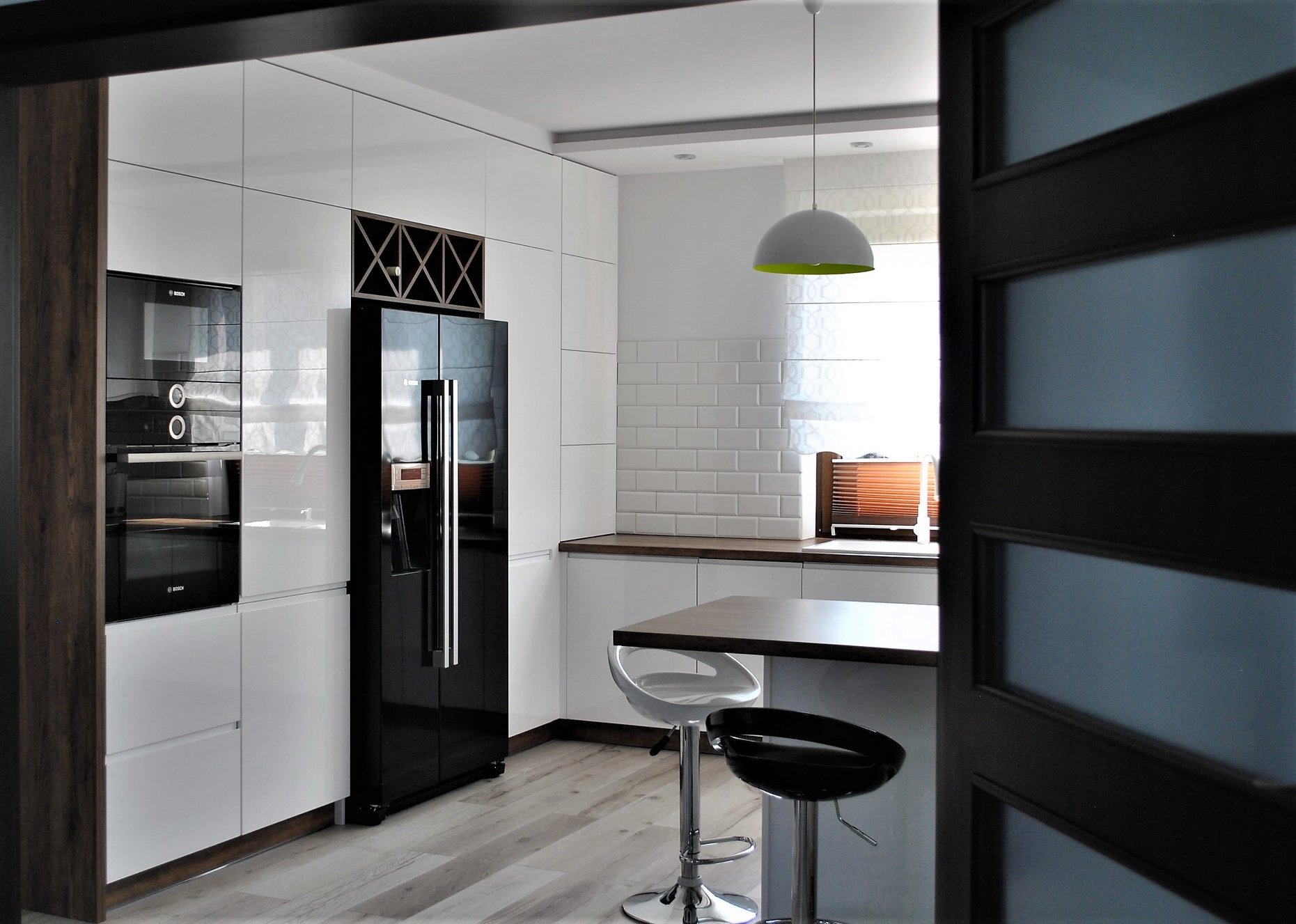 Nowoczesna kuchnia z białymi, błyszczącymi frontami, czarnym sprzętem AGD i drewnianym blatem, widziana z perspektywy otwartych drzwi.