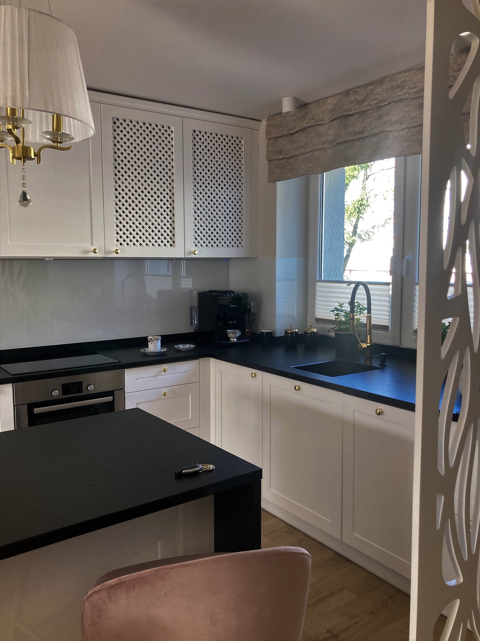 Nowoczesna kuchnia z białymi szafkami z ażurowymi frontami, czarnym blatem i wyspą, złotą armaturą, designerską lampą i dekoracyjną ścianką działową.
