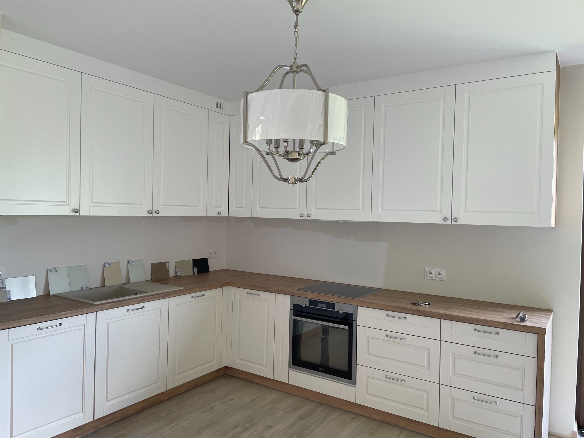 Klasyczna biała kuchnia z drewnianym blatem, widoczne próbki kolorów przy zlewie, ozdobny żyrandol, zabudowa pod sufit.