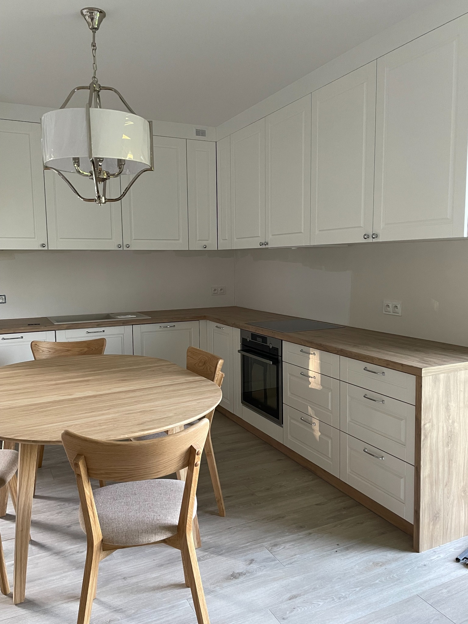 Jasna kuchnia z białymi szafkami, drewnianym blatem i okrągłym stołem z krzesłami w stylu skandynawskim, oświetlona lampą wiszącą z abażurem.