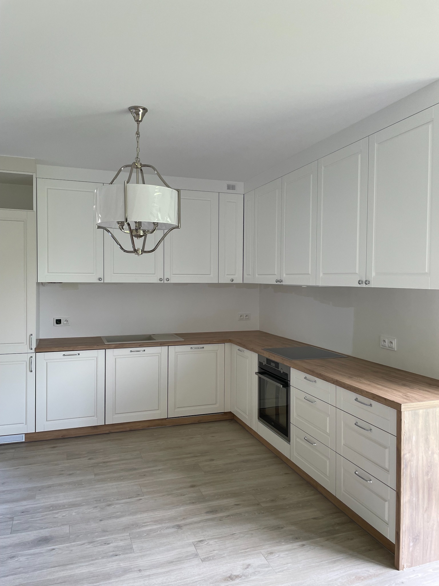 Jasna kuchnia w kształcie litery L z białymi frontami szafek i drewnianym blatem, oświetlona wiszącą lampą w stylu industrialnym.