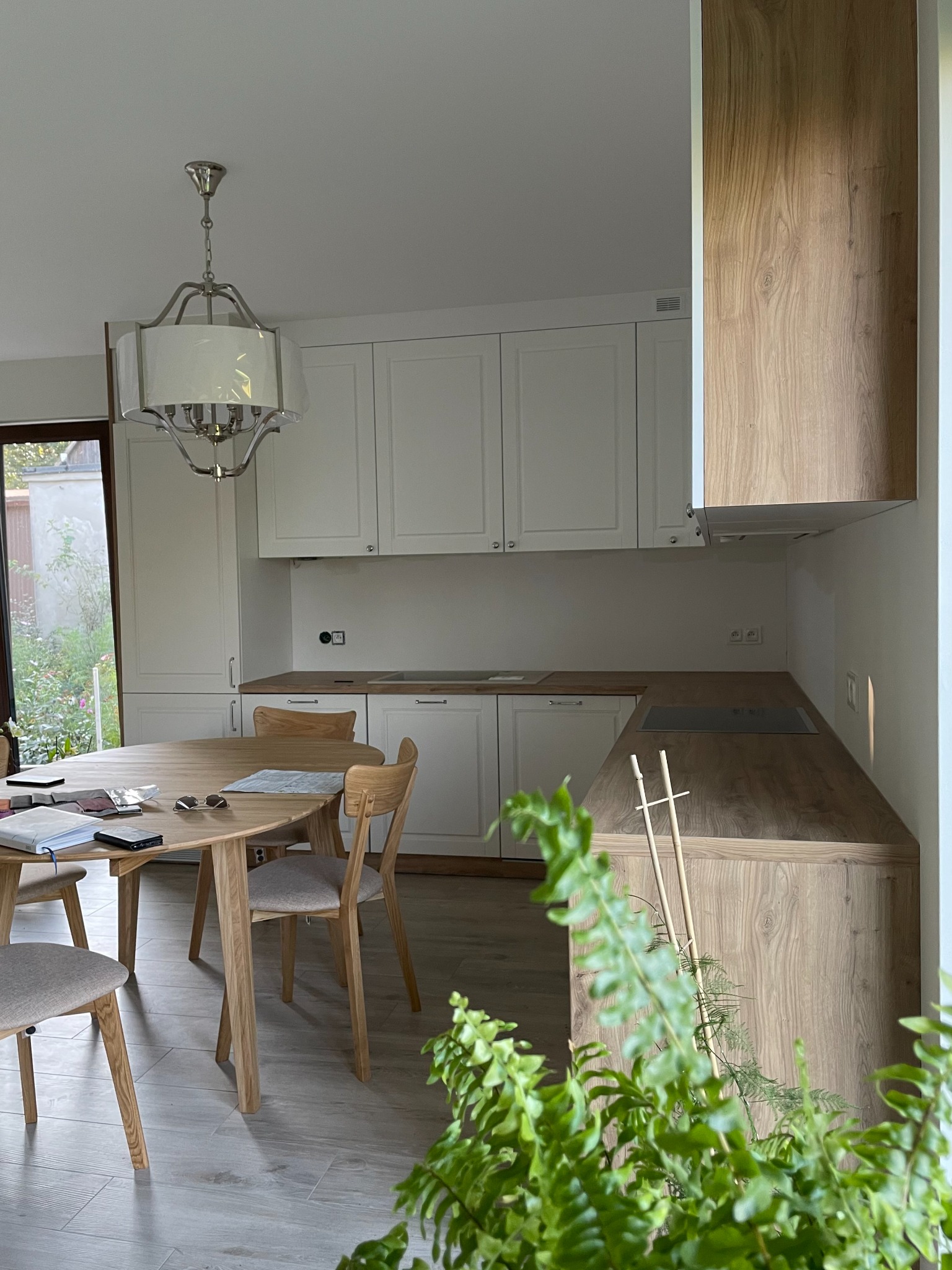 Jasna kuchnia z białymi szafkami i drewnianym blatem, okrągły stół z krzesłami, widok zza paprotki w doniczce.