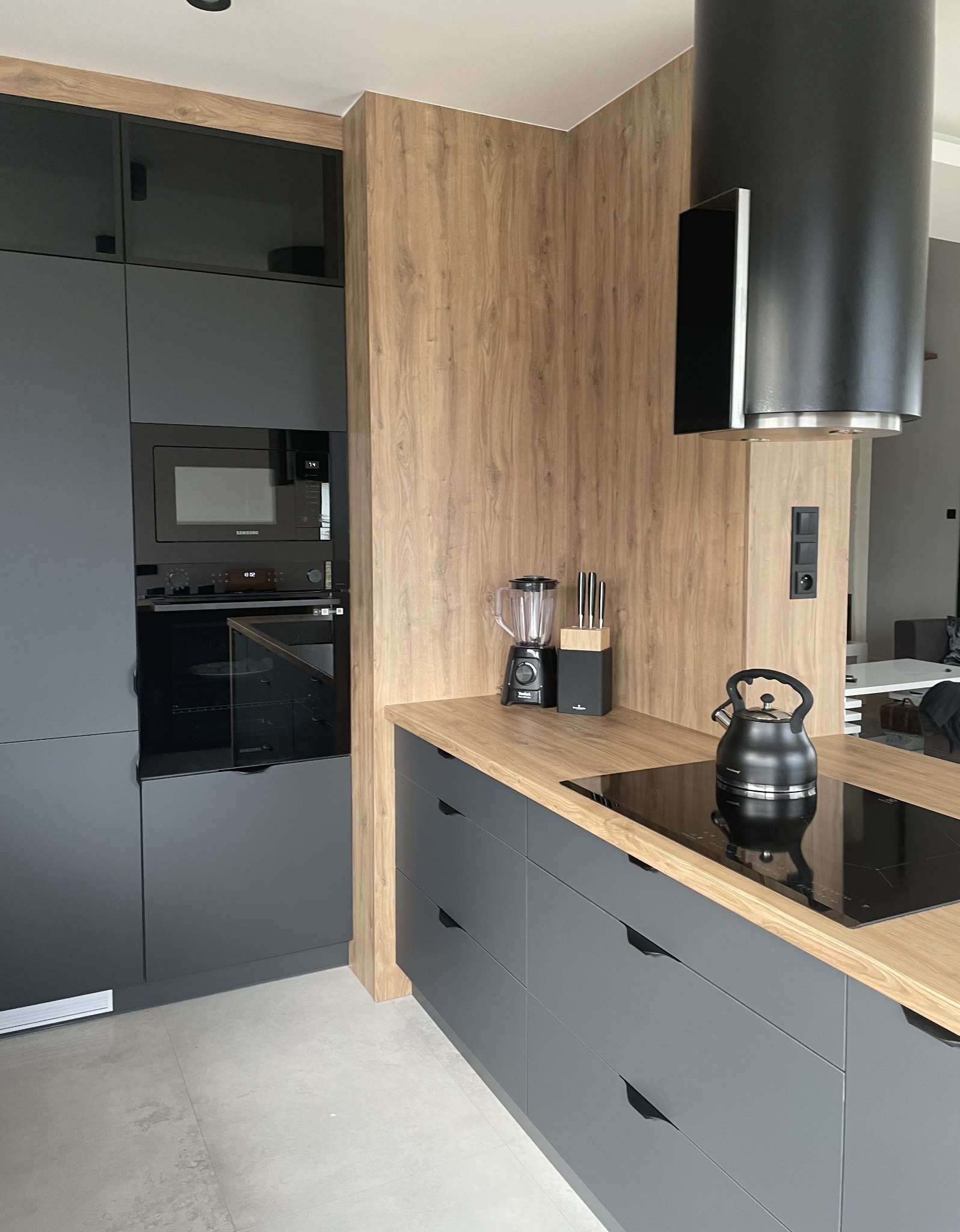Nowoczesna kuchnia: szare, matowe fronty szafek z drewnianym blatem, czarny czajnik na płycie indukcyjnej, blender i nóż w bloku na blacie, czarny okap cylindryczny.