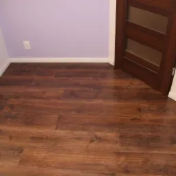 Ułożona podłoga z paneli imitujących drewno w odcieniach brązu, widoczna ściana pomalowana na fioletowo z gniazdkiem elektrycznym, fragment grzejnika, drzwi z przeszkleniami i fragment ściany...