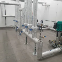 Rzędy izolowanych rur w pomieszczeniu technicznym, z widocznymi zaworami, manometrem i pompą obiegową z zielonym silnikiem, na podłodze z płytek.