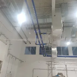 Wnętrze hali przemysłowej z widoczną instalacją wentylacyjną, niebieskie rury oraz srebrne kanały wentylacyjne pod sufitem, w tle okna i elementy konstrukcyjne budynku.