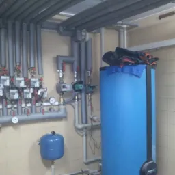 Instalacja CO z niebieskim zbiornikiem buforowym w kotłowni w Ochabach Małych. Rury i liczniki ciepła zamontowane na ścianie z beżowymi płytkami.