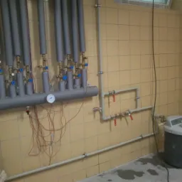 Szara instalacja CO z rurami, zaworami i manometrami na beżowej ścianie z płytek. W tle drabina i zbiornik z kubkiem na górze. Widok na instalację w kotłowni.