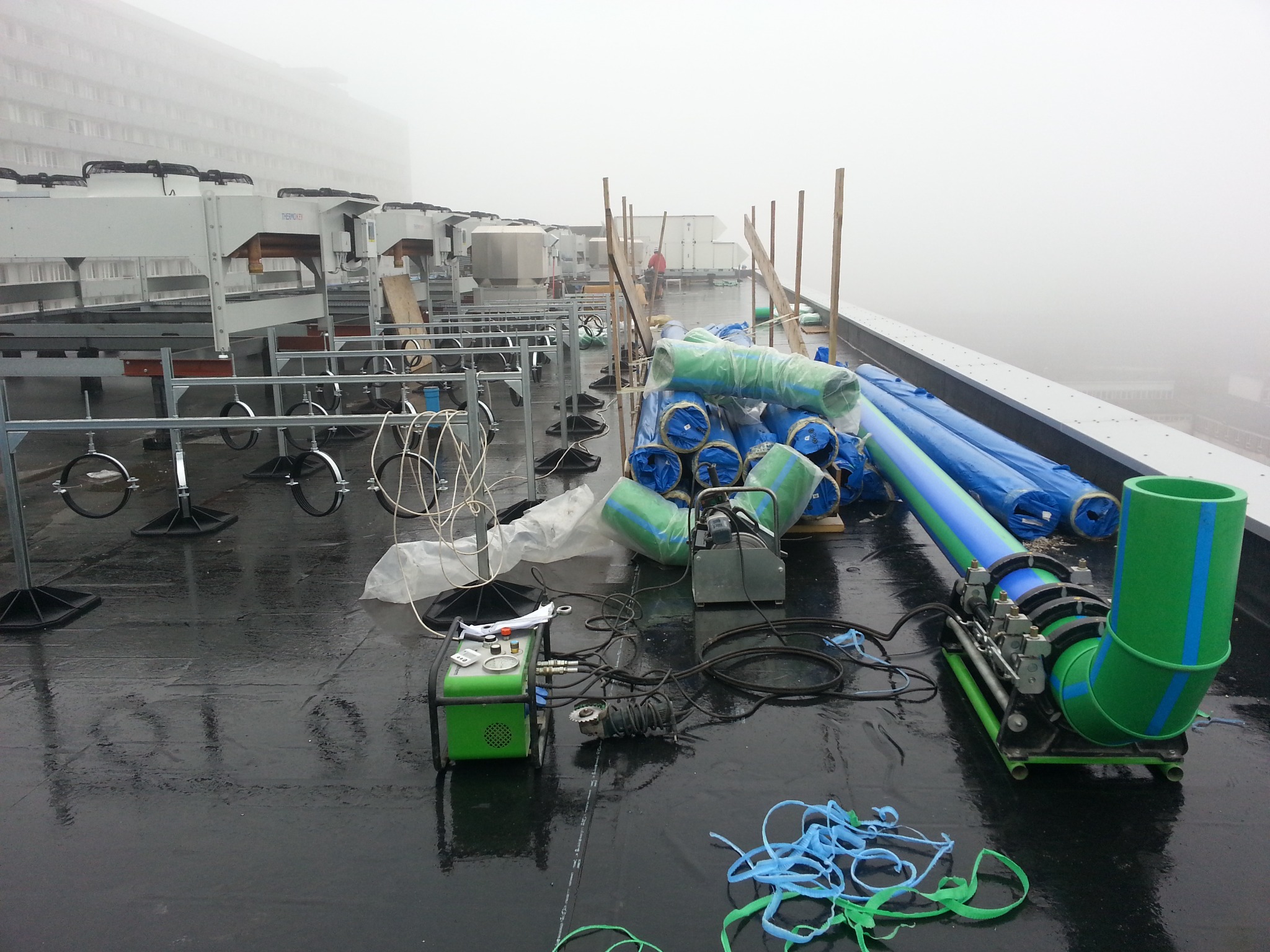 Montaż rur wentylacyjnych na dachu budynku w mglisty dzień, widoczny sprzęt do łączenia rur i zwinięte rury w niebiesko-zielonej folii.