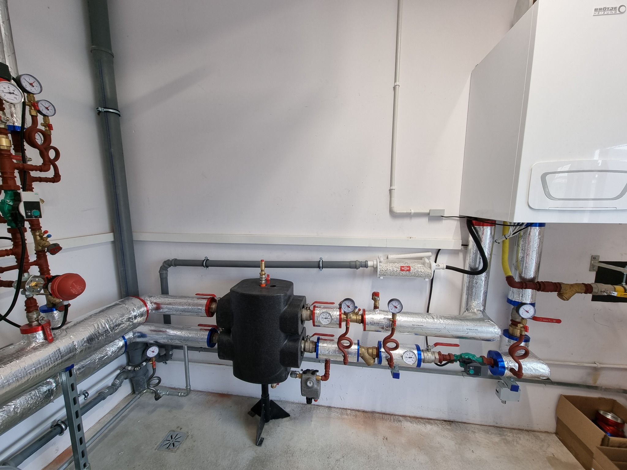 Kocioł gazowy z podłączeniami hydraulicznymi i izolowanymi rurami w pomieszczeniu technicznym, widoczne manometry i zawory, czyste białe ściany i szary beton na podłodze.