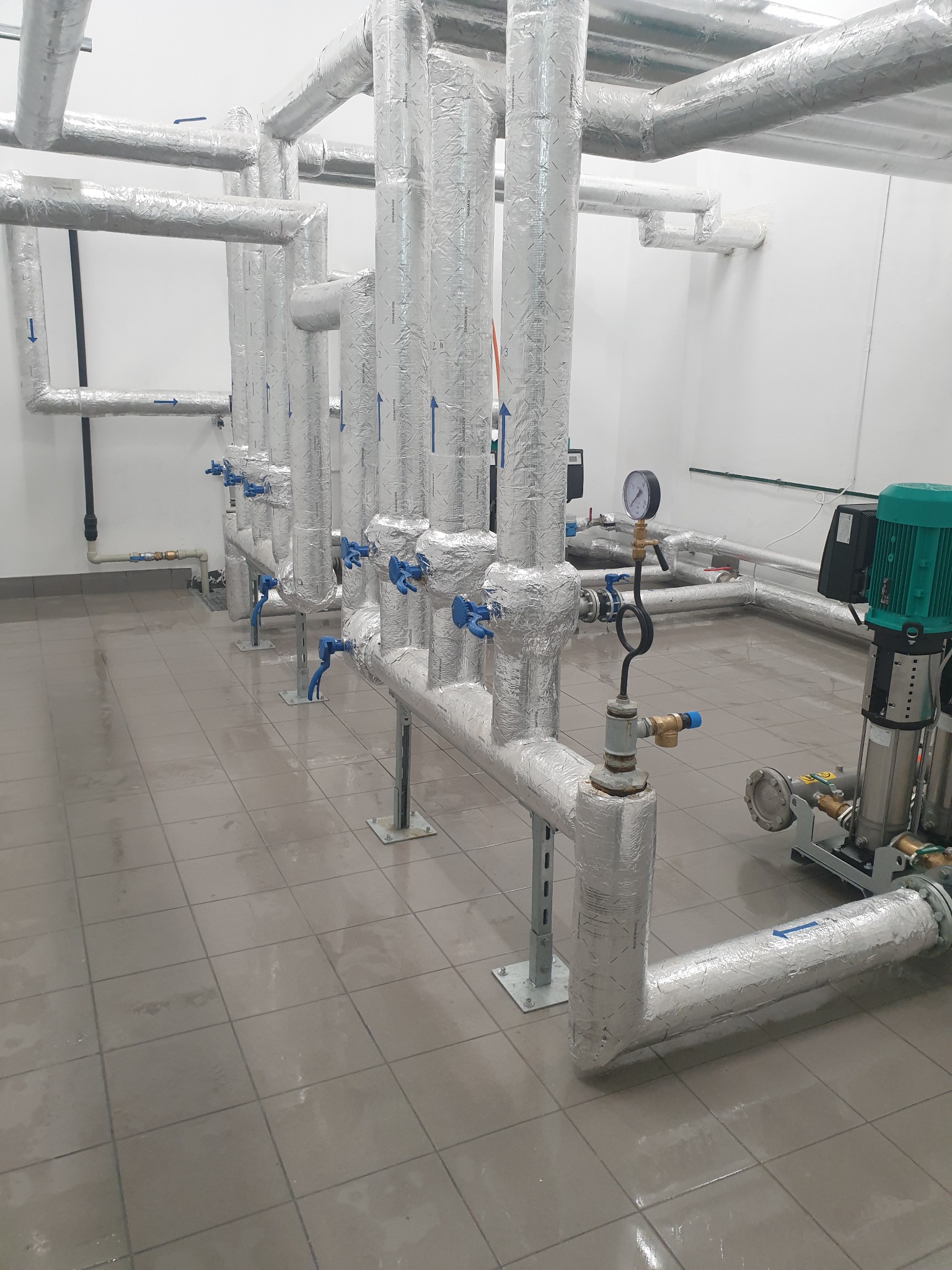 Rzędy izolowanych rur w pomieszczeniu technicznym, z widocznymi zaworami, manometrem i pompą obiegową z zielonym silnikiem, na podłodze z płytek.