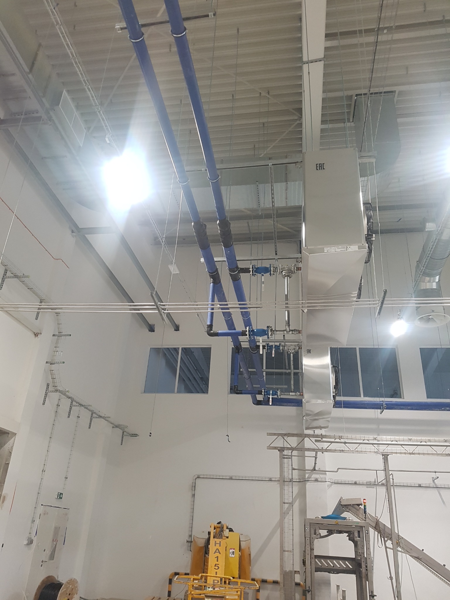 Wnętrze hali przemysłowej z widoczną instalacją wentylacyjną, niebieskie rury oraz srebrne kanały wentylacyjne pod sufitem, w tle okna i elementy konstrukcyjne budynku.