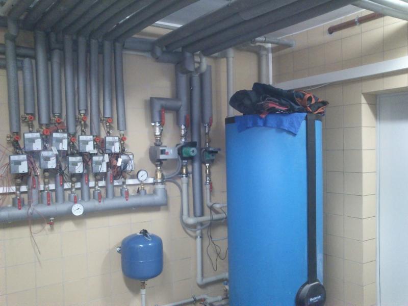 Instalacja CO z niebieskim zbiornikiem buforowym w kotłowni w Ochabach Małych. Rury i liczniki ciepła zamontowane na ścianie z beżowymi płytkami.