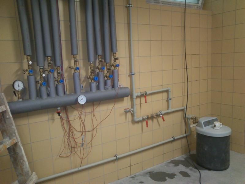 Szara instalacja CO z rurami, zaworami i manometrami na beżowej ścianie z płytek. W tle drabina i zbiornik z kubkiem na górze. Widok na instalację w kotłowni.