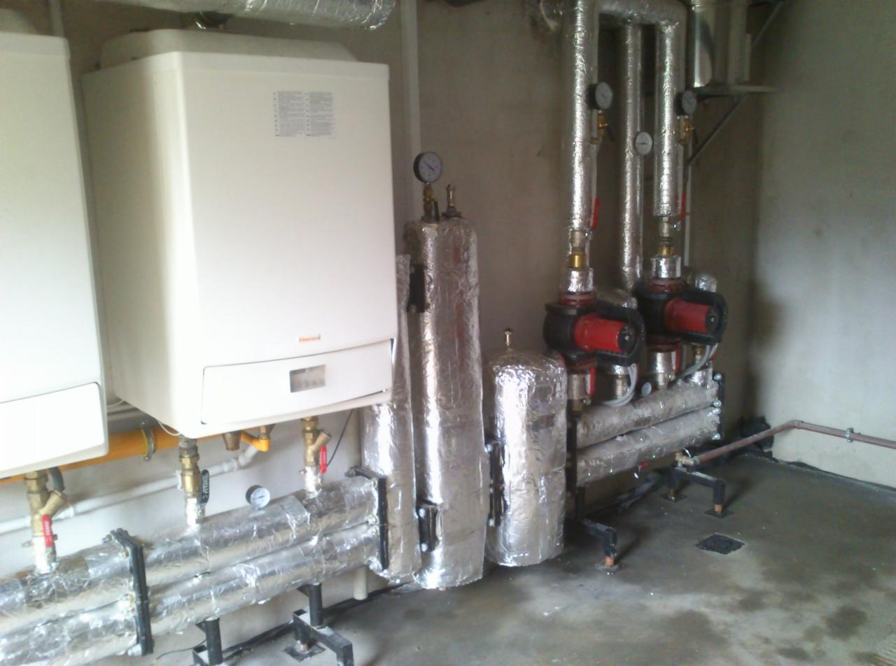 Dwa białe piece gazowe wiszące na ścianie, z widocznymi rurami w izolacji, pompami obiegowymi i zbiornikami wyrównawczymi w pomieszczeniu technicznym.