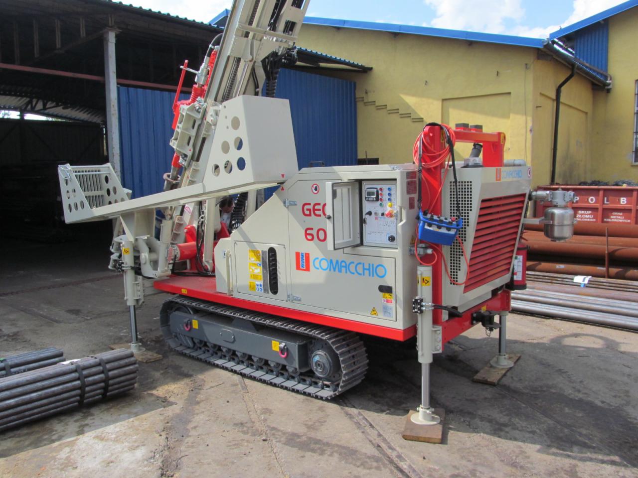 Wiertnica Comacchio GEO 60 na gąsienicach, z wysuniętymi podporami, przygotowana do odwiertów geotechnicznych, na tle budynku z blachy falistej i żółtej ściany.