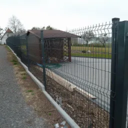 Nowoczesne ogrodzenie panelowe w kolorze grafitowym, z podmurówką betonową, otaczające posesję z budynkiem i altaną w tle, widok z boku na żwirową ścieżkę.