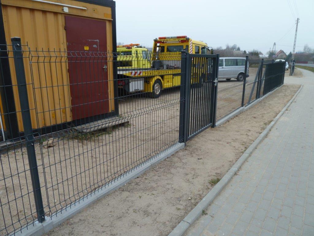 Ciemnoszare ogrodzenie panelowe z furtką na betonowej podmurówce, z żółtymi pojazdami w tle, ustawione wzdłuż chodnika z kostki brukowej.
