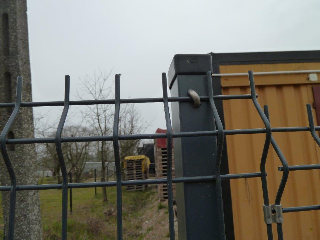 Fragment grafitowego ogrodzenia panelowego na tle zieleni i budynku. Widoczne detale mocowania i struktura siatki, podkreślone pochmurnym oświetleniem.