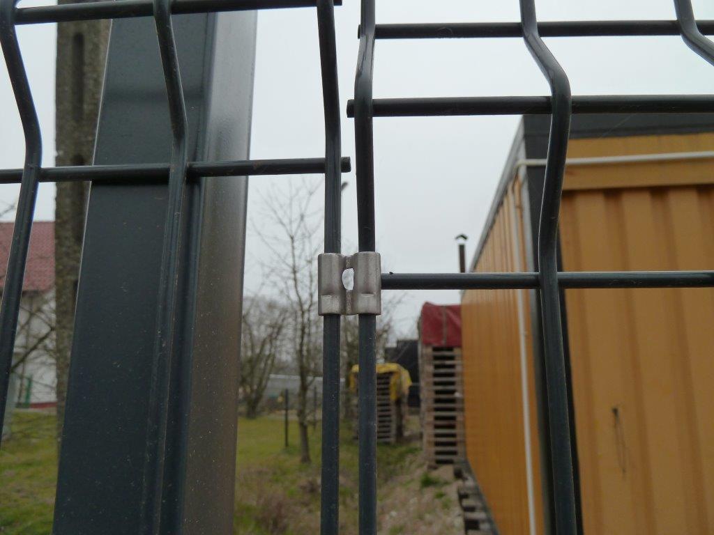 Szczegółowy widok z bliska na metalowe ogrodzenie panelowe z łącznikiem, ukazujący jego solidną konstrukcję i precyzyjne połączenia na tle zieleni i budynku.